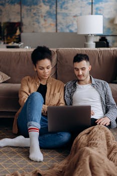 A young couple sitting together on the floor, focused on a laptop in a cozy home setting.